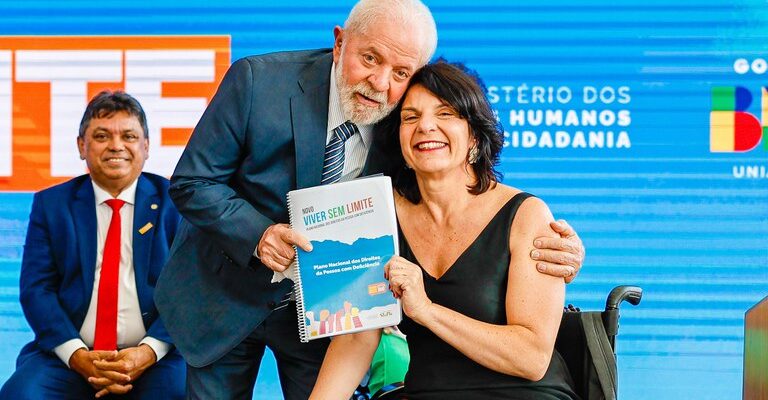 O presidente Lula está ao lado de uma mulher em cadeira de rodas, segurando um documento sobre direitos da pessoa com deficiência. Ao fundo, há um painel do Ministério dos Direitos Humanos e Cidadania.