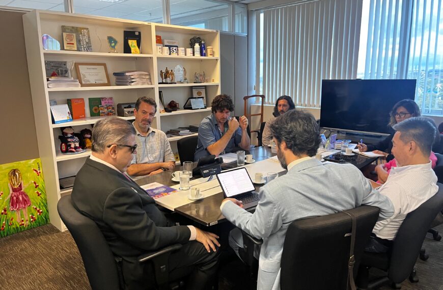 Descrição: Sala de reunião com sete pessoas sentadas ao redor de uma mesa retangular. A mesa está coberta com xícaras de café, copos de água e alguns papéis. Um dos homens está usando um laptop. Ao fundo, há uma estante com prateleiras cheias de livros, troféus e objetos decorativos. Na parede à esquerda, há uma pintura de uma menina de vestido rosa em um campo de flores. As janelas ao fundo têm persianas verticais, permitindo a entrada de luz natural. Uma televisão grande está posicionada ao lado das janelas. As pessoas parecem estar em uma discussão ou reunião de trabalho.