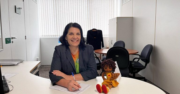 Mulher de cabelos curtos e escuros, vestindo blazer cinza, está sorrindo e escrevendo em uma mesa branca de escritório. Ao lado dela, há uma boneca de crochê. O ambiente tem uma mesa redonda, cadeiras pretas e um armário branco, com luz entrando pelas persianas.