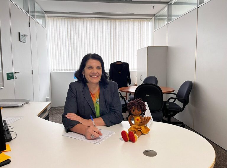 Mulher de cabelos curtos e escuros, vestindo blazer cinza, está sorrindo e escrevendo em uma mesa branca de escritório. Ao lado dela, há uma boneca de crochê. O ambiente tem uma mesa redonda, cadeiras pretas e um armário branco, com luz entrando pelas persianas.