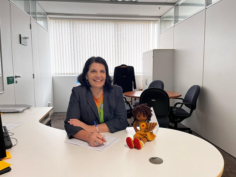 Mulher de cabelos curtos e escuros, vestindo blazer cinza, está sorrindo e escrevendo em uma mesa branca de escritório. Ao lado dela, há uma boneca de crochê. O ambiente tem uma mesa redonda, cadeiras pretas e um armário branco, com luz entrando pelas persianas.