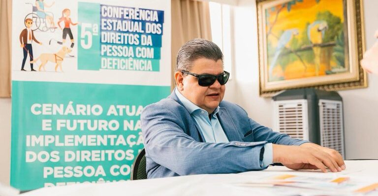 Homem de óculos escuros em uma conferência sobre direitos das pessoas com deficiência, com as mãos em um livro fazendo leitura em Braille. Ao fundo, um banner e uma pintura de araras na parede.