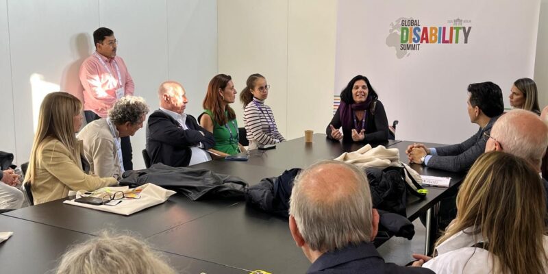 Um grupo de pessoas está em uma reunião ao redor de uma mesa, com uma mulher liderando a discussão. Ao fundo, há um banner do "Global Disability Summit". A atmosfera é formal e atenta.