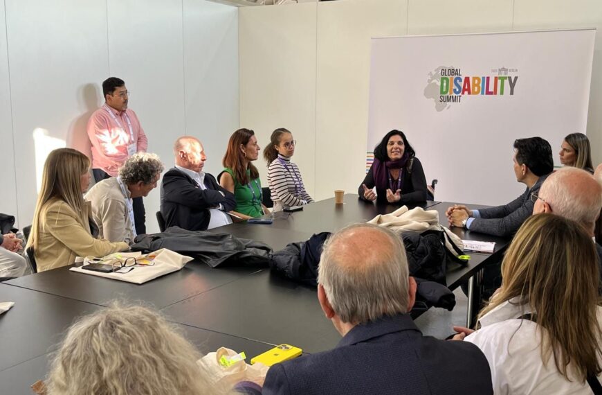 Um grupo de pessoas está em uma reunião ao redor de uma mesa, com uma mulher liderando a discussão. Ao fundo, há um banner do "Global Disability Summit". A atmosfera é formal e atenta.