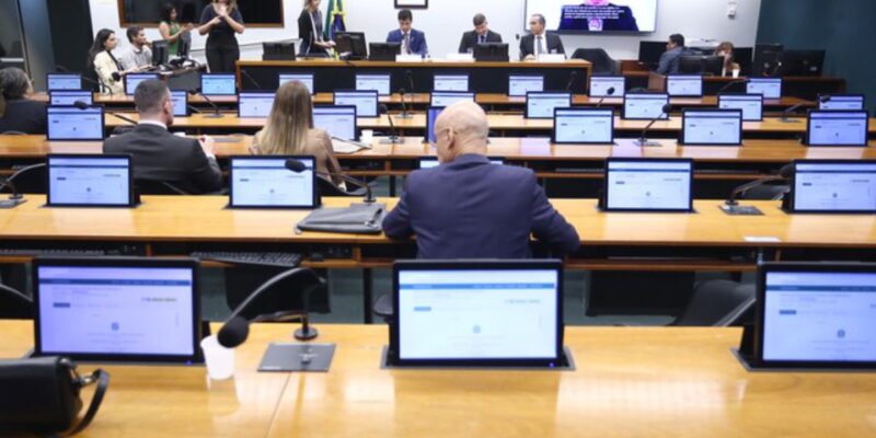 Ambiente de reunião parlamentar com pessoas sentadas, monitores nas mesas e uma videoconferência sendo exibida ao fundo.