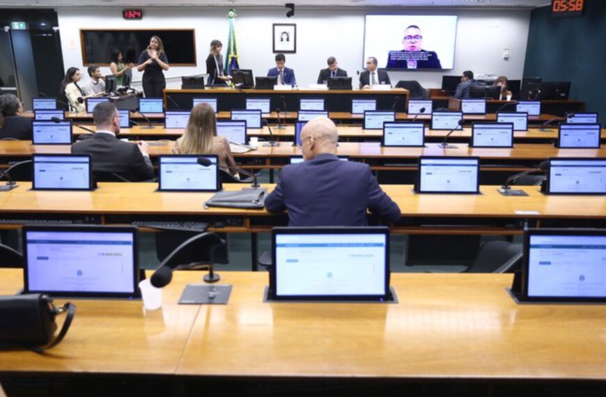 Sala de reunião parlamentar com computadores, pessoas em trajes formais e intérprete de Libras.