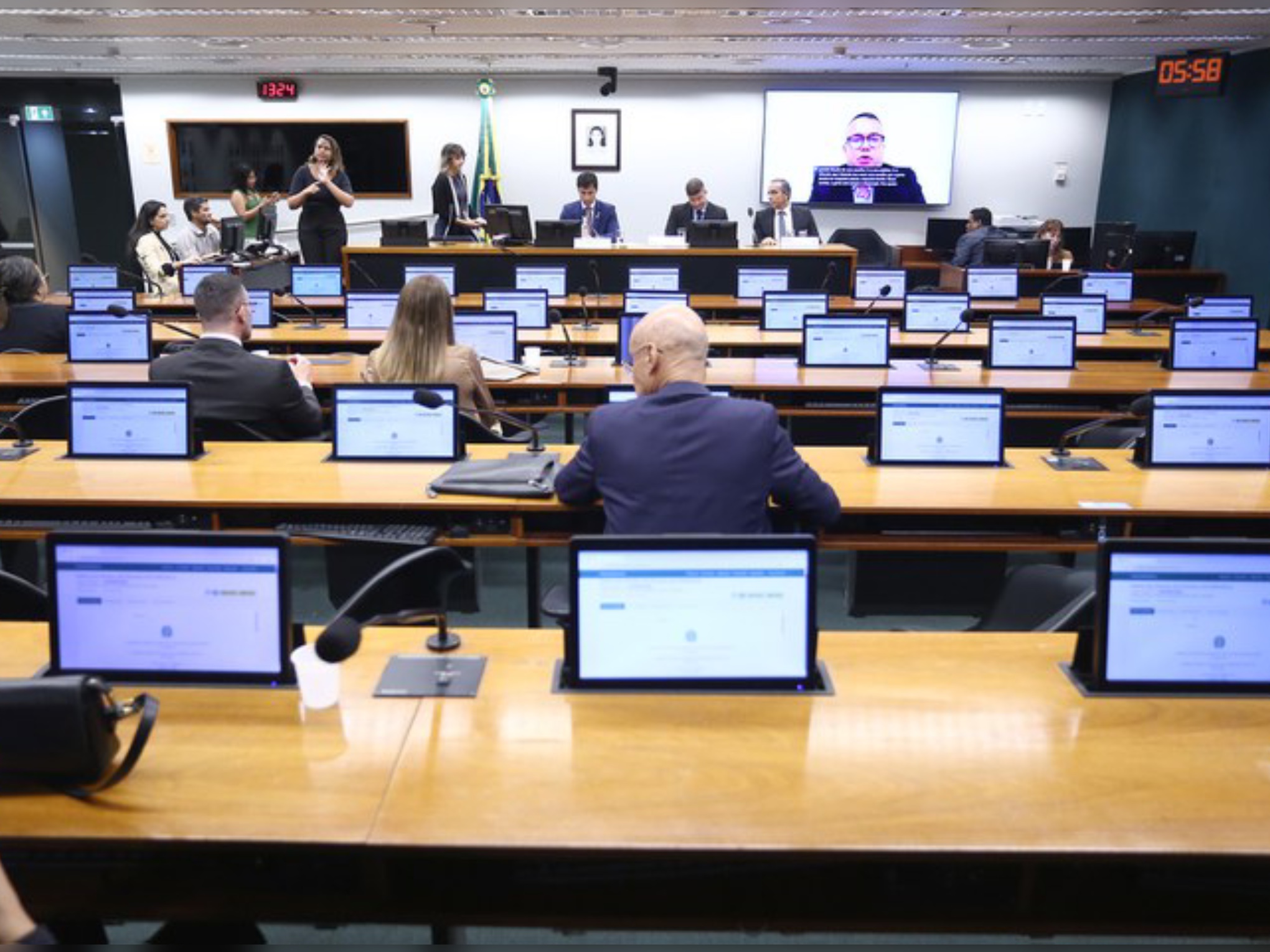 Ambiente de reunião parlamentar com pessoas sentadas, monitores nas mesas e uma videoconferência sendo exibida ao fundo.