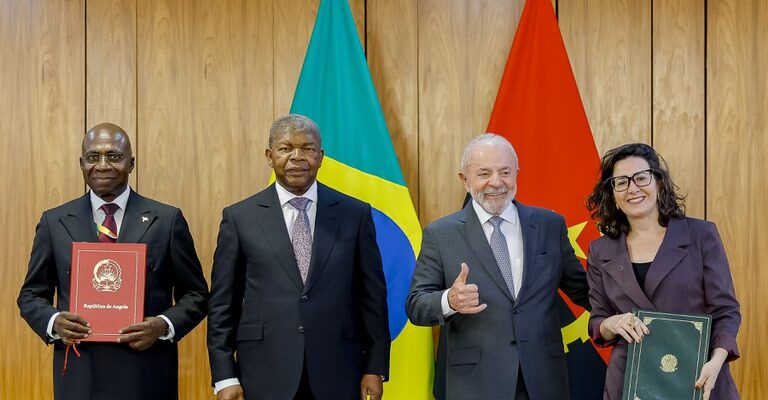 Quatro pessoas estão em pé diante das bandeiras do Brasil e de Angola, participando de um evento oficial. Entre elas está o presidente Lula, que aparece sorrindo e fazendo sinal de positivo com a mão. Ao lado dele há uma mulher segurando uma pasta verde.