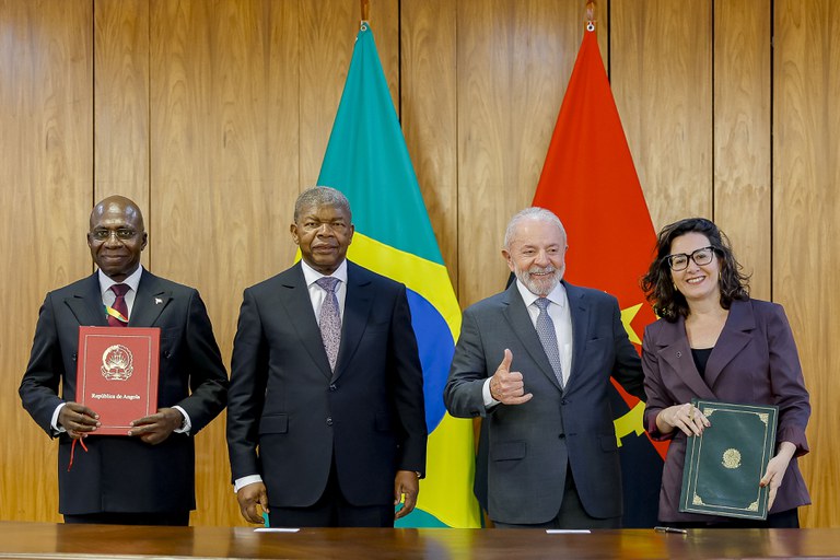 Quatro pessoas estão em pé diante das bandeiras do Brasil e de Angola, participando de um evento oficial. Entre elas está o presidente Lula, que aparece sorrindo e fazendo sinal de positivo com a mão. Ao lado dele há uma mulher segurando uma pasta verde.