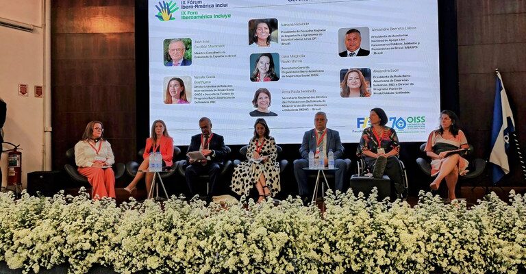Uma conferência com sete pessoas no palco, um painel de LED ao fundo exibindo informações do "IX Fórum Iberoamérica Inclui" e uma decoração de flores brancas na frente do palco.i