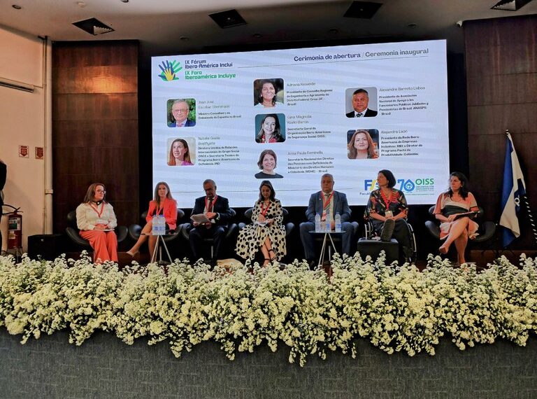 Uma conferência com sete pessoas no palco, um painel de LED ao fundo exibindo informações do "IX Fórum Iberoamérica Inclui" e uma decoração de flores brancas na frente do palco.i