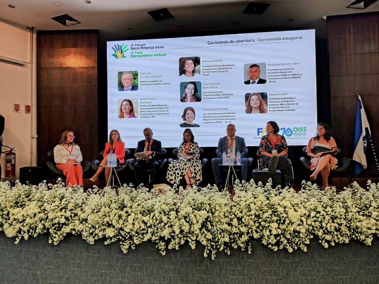 Uma conferência com sete pessoas no palco, um painel de LED ao fundo exibindo informações do "IX Fórum Iberoamérica Inclui" e uma decoração de flores brancas na frente do palco.i