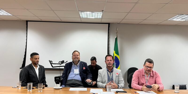 Quatro homens sentados atrás de uma mesa em uma sala de reunião, com copos de água e papéis à frente. Ao fundo, há uma tela de projeção e uma bandeira do Brasil. O ambiente é formal e parece ser uma reunião ou conferência.
