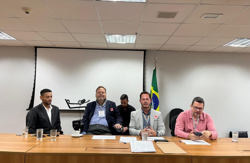 Quatro homens sentados atrás de uma mesa em uma sala de reunião, com copos de água e papéis à frente. Ao fundo, há uma tela de projeção e uma bandeira do Brasil. O ambiente é formal e parece ser uma reunião ou conferência.