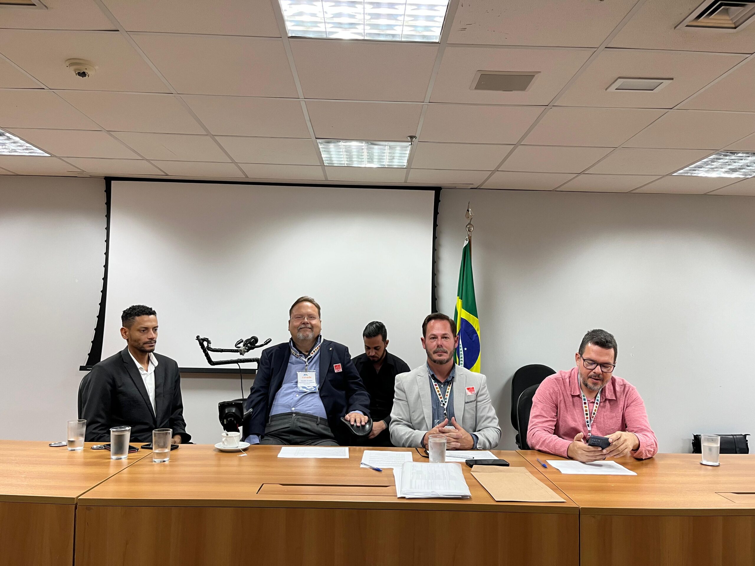 Quatro homens sentados atrás de uma mesa em uma sala de reunião, com copos de água e papéis à frente. Ao fundo, há uma tela de projeção e uma bandeira do Brasil. O ambiente é formal e parece ser uma reunião ou conferência.