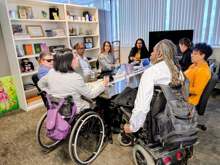 Reunião em uma sala de conferências com oito pessoas, incluindo duas em cadeiras de rodas, ao redor de uma mesa. A sala tem uma estante com livros e prêmios, e as pessoas estão discutindo ou apresentando, com copos de água na mesa.