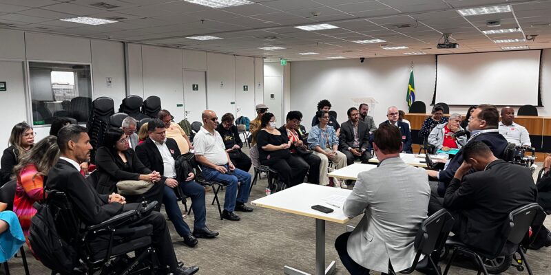 Reunião em uma sala corporativa, com cerca de 30 pessoas, incluindo algumas em cadeiras de rodas, sentadas em círculo e participando de uma discussão séria. Há uma mesa de tribunal e uma bandeira do Brasil ao fundo.