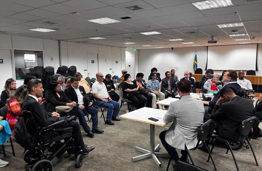 Reunião em uma sala corporativa, com cerca de 30 pessoas, incluindo algumas em cadeiras de rodas, sentadas em círculo e participando de uma discussão séria. Há uma mesa de tribunal e uma bandeira do Brasil ao fundo.