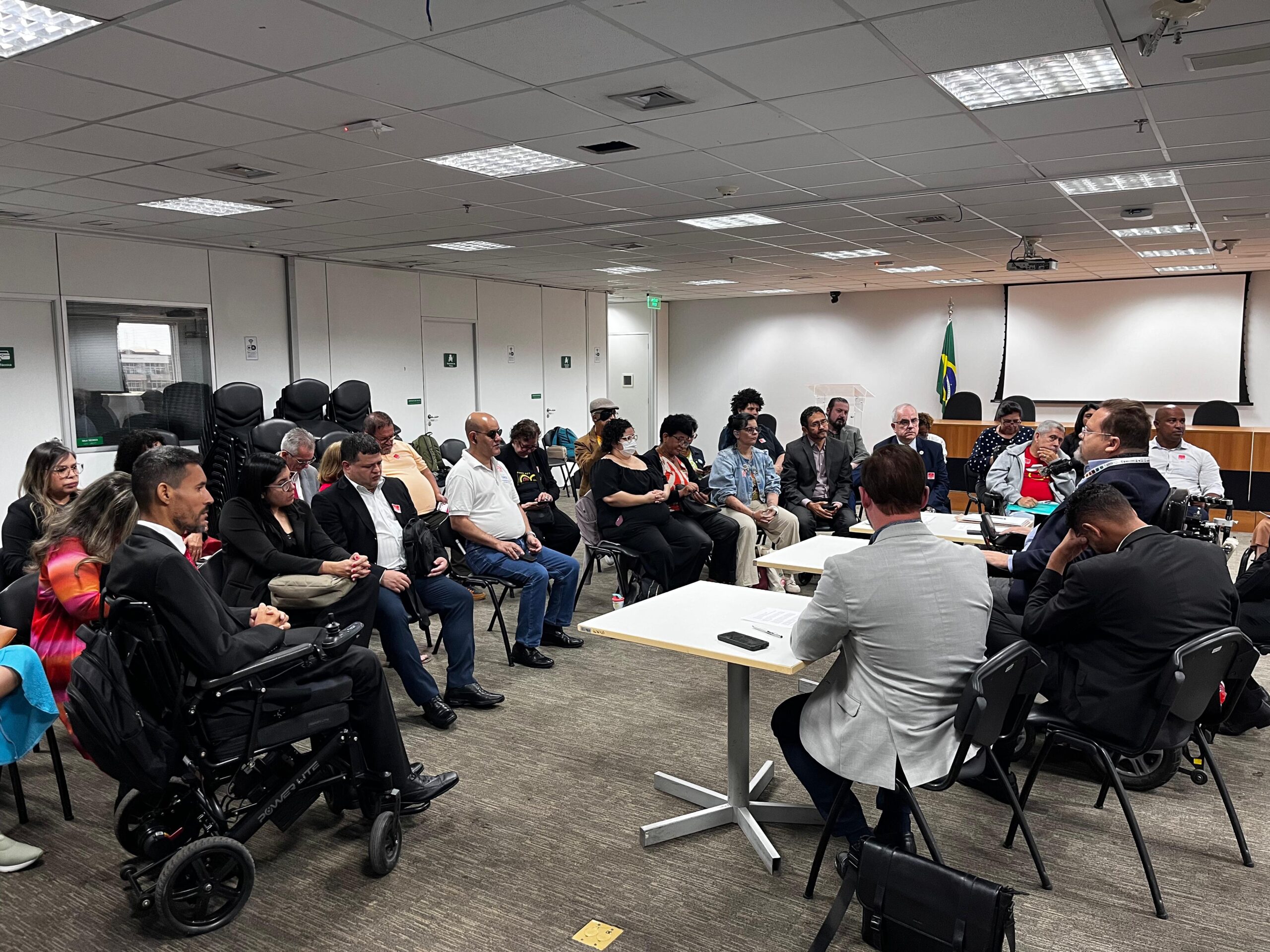 Reunião em uma sala corporativa, com cerca de 30 pessoas, incluindo algumas em cadeiras de rodas, sentadas em círculo e participando de uma discussão séria. Há uma mesa de tribunal e uma bandeira do Brasil ao fundo.