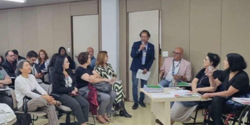 Reunião formal com cerca de 30 pessoas, onde a maioria está sentada ouvindo uma apresentação feita por quatro pessoas à frente, incluindo uma mulher em cadeira de rodas. Um homem fala ao microfone. O ambiente é iluminado e organizado, sugerindo um evento profissional ou acadêmico.