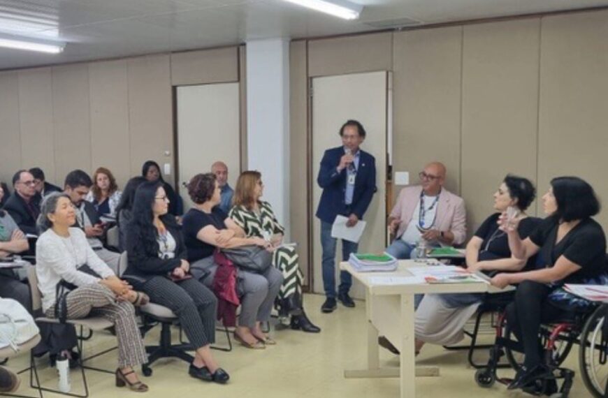 Reunião com pessoas sentadas, um homem fala ao microfone e há uma mulher em cadeira de rodas.