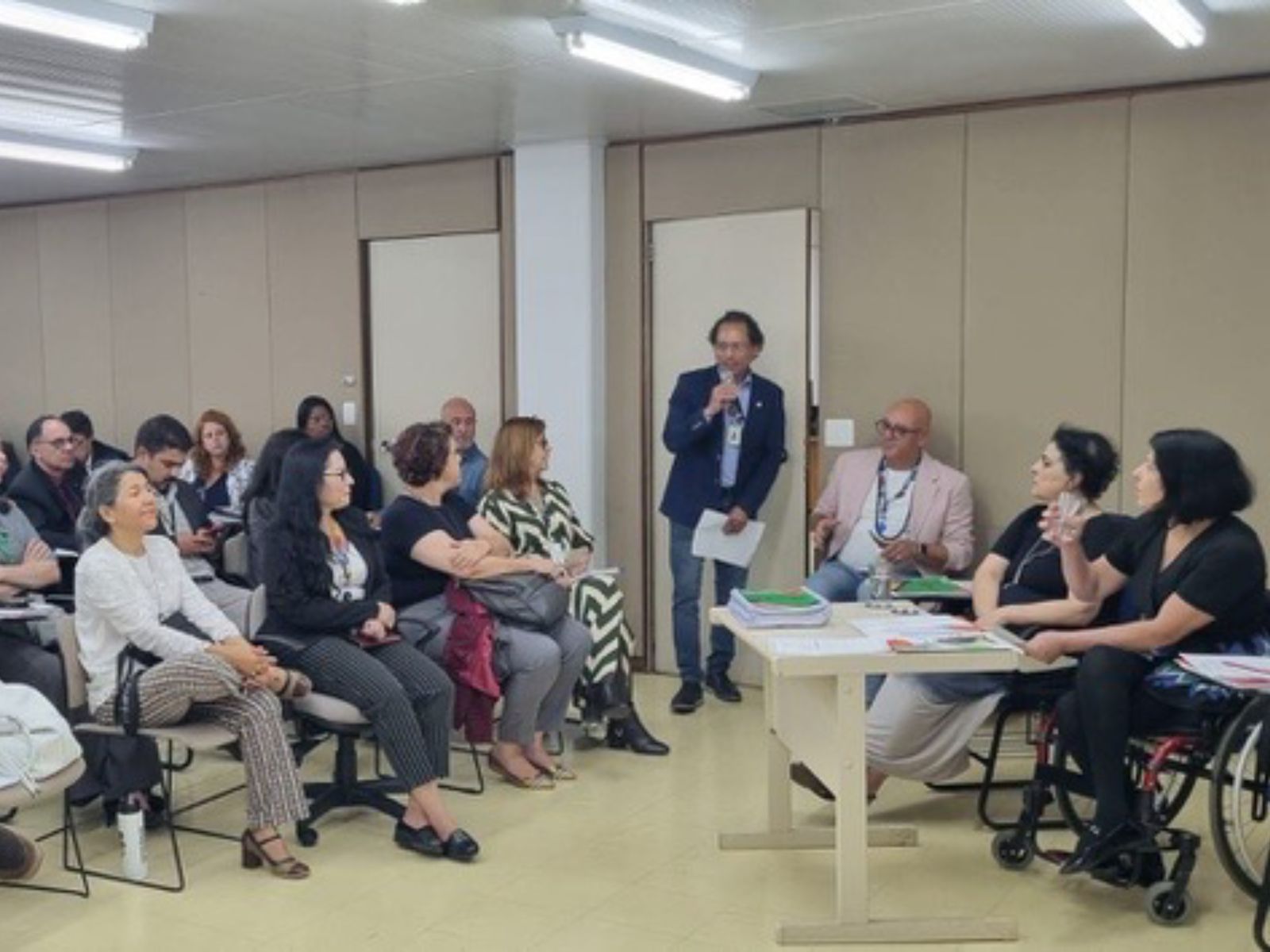 Reunião formal com cerca de 30 pessoas, onde a maioria está sentada ouvindo uma apresentação feita por quatro pessoas à frente, incluindo uma mulher em cadeira de rodas. Um homem fala ao microfone. O ambiente é iluminado e organizado, sugerindo um evento profissional ou acadêmico.
