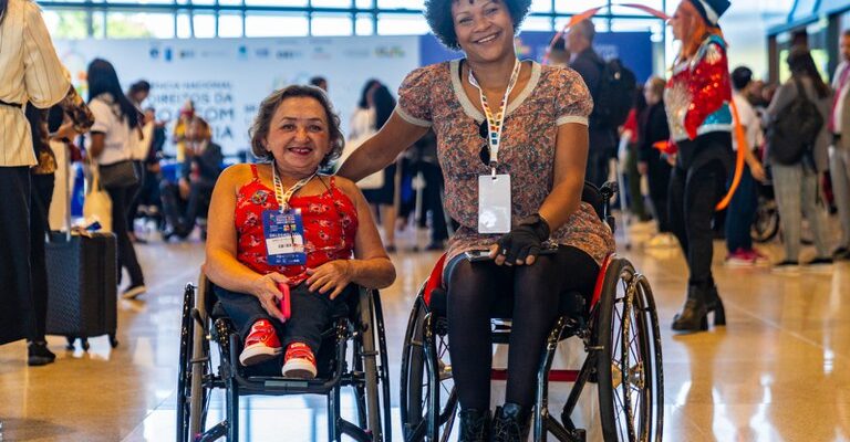 Duas mulheres em cadeiras de rodas, sorrindo e usando crachás, estão lado a lado em um evento movimentado e inclusivo. Ao fundo, há várias pessoas e uma pessoa fantasiada, em um ambiente bem iluminado.