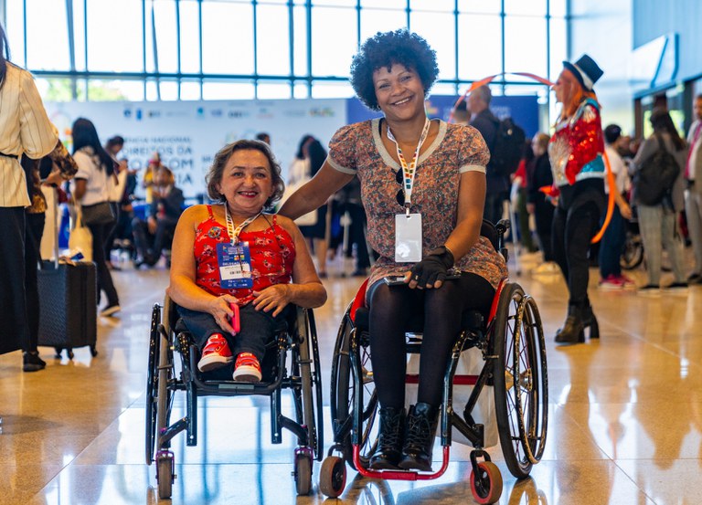 Duas mulheres em cadeiras de rodas, sorrindo e usando crachás, estão lado a lado em um evento movimentado e inclusivo. Ao fundo, há várias pessoas e uma pessoa fantasiada, em um ambiente bem iluminado.