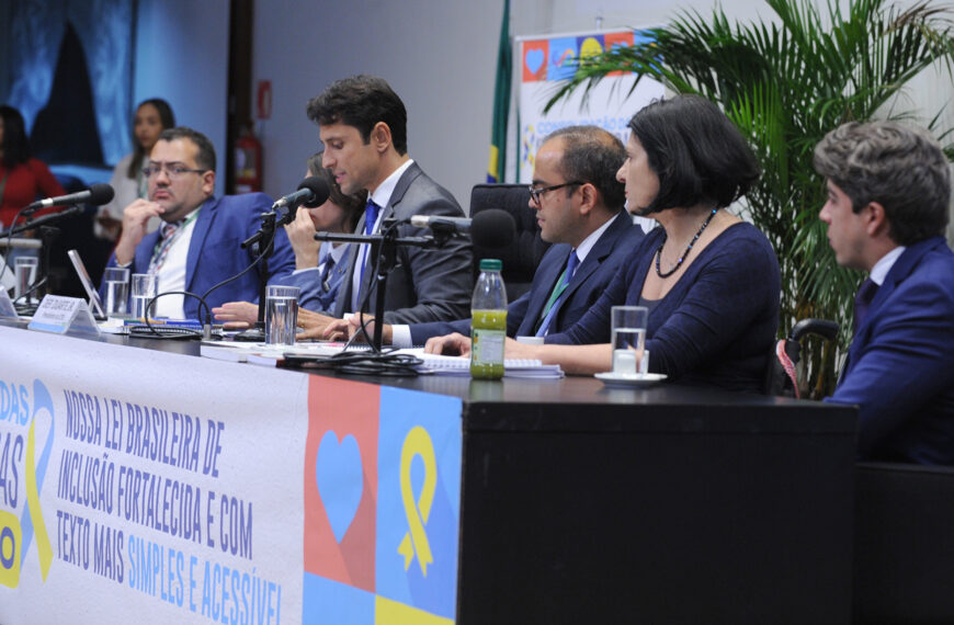 Seis pessoas estão sentadas em uma mesa de conferência, todas vestidas formalmente. Um homem no centro fala ao microfone, enquanto os outros, incluindo uma mulher e homens de terno, participam da discussão.