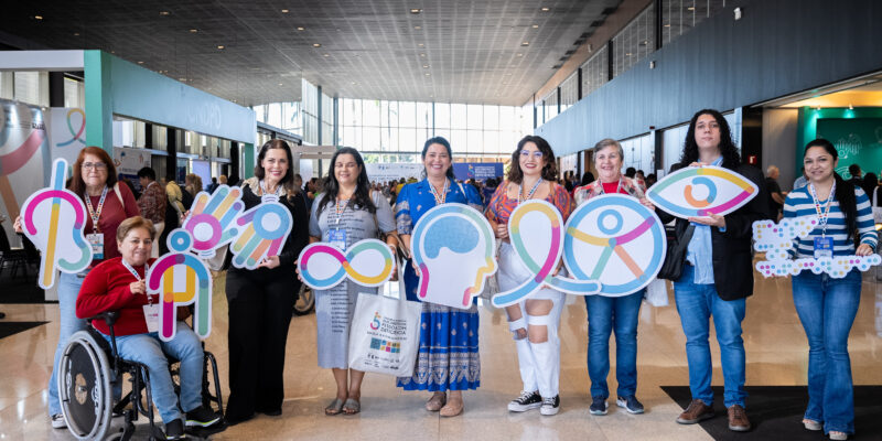 Nove pessoas em um evento interno seguram placas com símbolos de diferentes tipos de deficiência. Uma delas está em cadeira de rodas. Todos usam crachás e sorriem, em um espaço amplo com janelas grandes e estandes ao fundo.