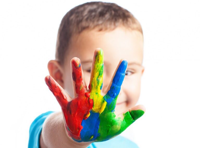 Criança sorridente com a mão levantada em direção à câmera, coberta de tinta colorida. As cores na mão são vermelho, amarelo, azul e verde, criando um efeito vibrante. O fundo é branco, destacando ainda mais as cores vivas na mão da criança. A criança está usando uma camiseta azul clara.