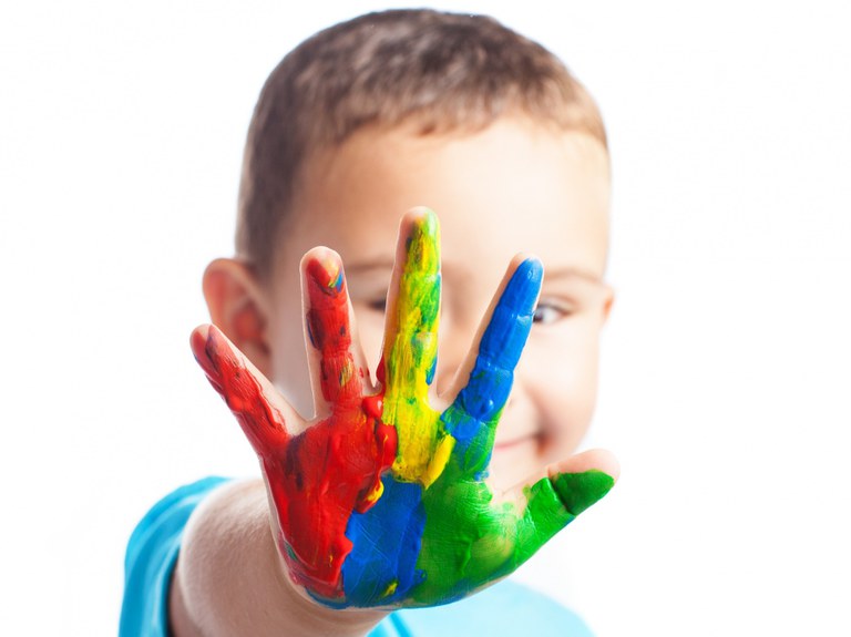 Criança sorridente com a mão levantada em direção à câmera, coberta de tinta colorida. As cores na mão são vermelho, amarelo, azul e verde, criando um efeito vibrante. O fundo é branco, destacando ainda mais as cores vivas na mão da criança. A criança está usando uma camiseta azul clara.