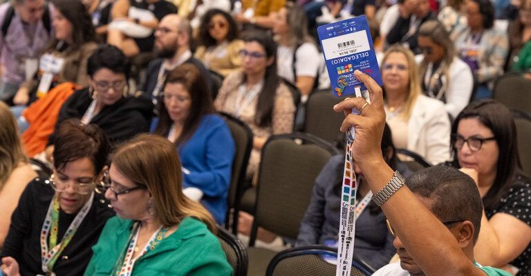 Uma conferência com várias pessoas sentadas, onde um homem levanta um crachá azul com a palavra "DELEGADO". As pessoas ao redor usam crachás e estão em um auditório.
