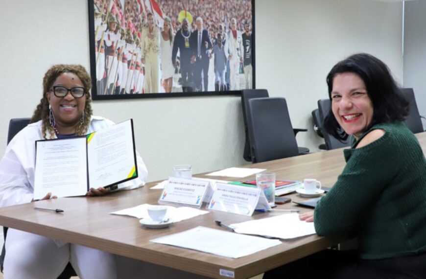 Duas mulheres sorrindo em reunião, uma segura documento aberto; mesa com papéis e xícaras.