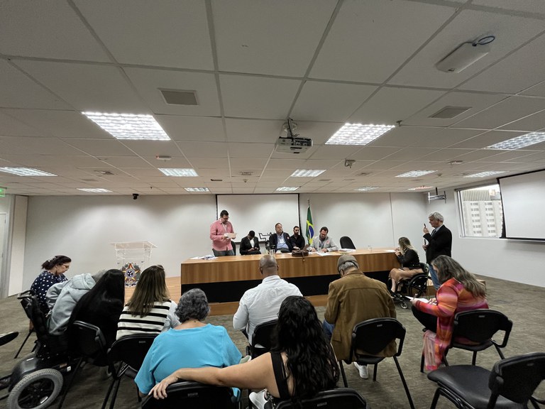 Reunião formal com pessoas sentadas, incluindo algumas em cadeiras de rodas, e uma mesa principal com cinco participantes. Ambiente institucional e organizado.ual