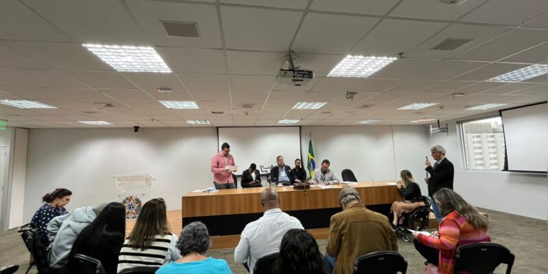 Reunião formal, pessoas sentadas ouvindo seis pessoas à mesa, bandeira do Brasil ao fundo.