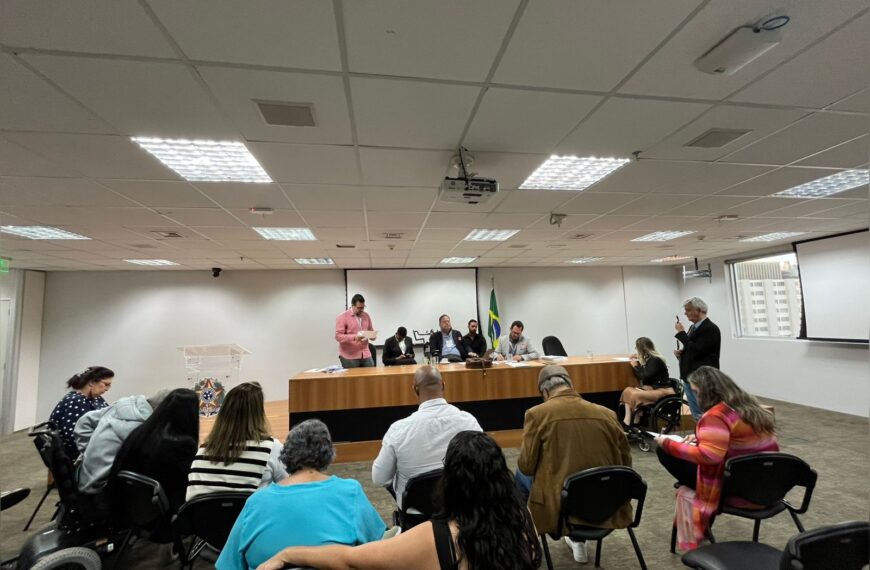Reunião formal com pessoas sentadas, algumas em cadeiras de rodas, e uma mesa principal com representantes. Ambiente institucional e organizado.