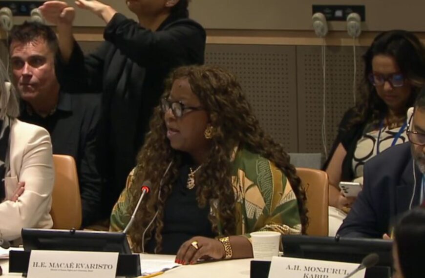 Mulher negra fala ao microfone em conferência, com intérprete de Libras ao fundo.