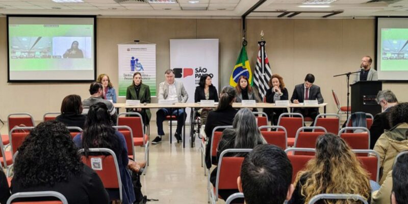 Mesa de debate formal com oito pessoas, público assistindo e bandeiras ao fundo.