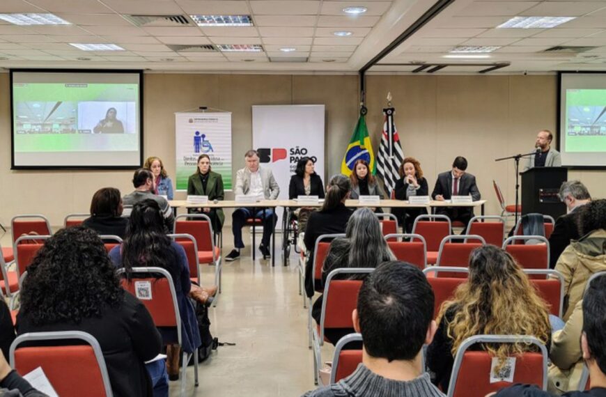 Mesa de debate formal com oito pessoas, público assistindo e bandeiras ao fundo.