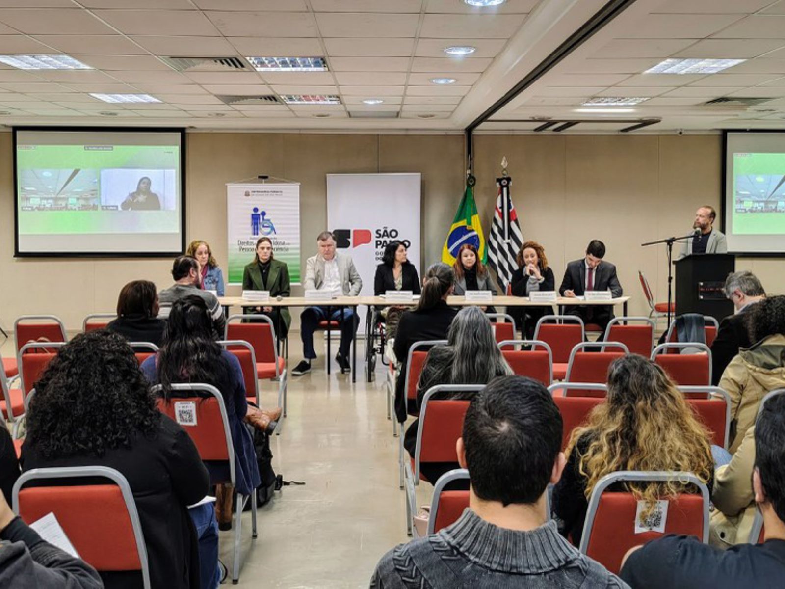 Mesa de debate formal com oito pessoas, público assistindo e bandeiras ao fundo.