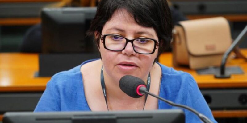 Mulher branca de cabelos curtos e óculos fala ao microfone em uma sala de reunião