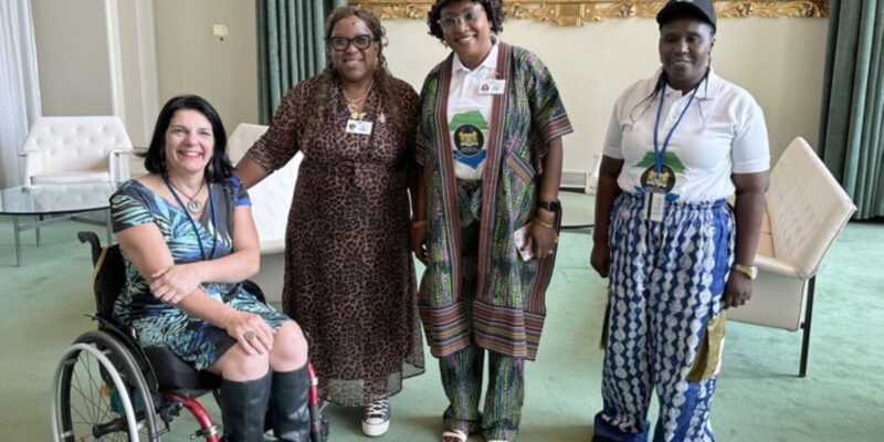 Quatro mulheres sorrindo, uma em cadeira de rodas, em ambiente formal com crachás.