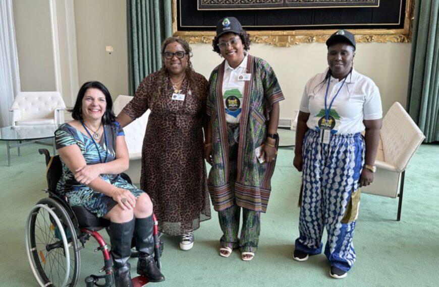 Quatro mulheres posam juntas em um ambiente elegante. Uma delas está em cadeira de rodas, duas usam roupas tradicionais africanas e crachás, e outra veste um vestido de oncinha. Todas parecem sorridentes e o local tem poltronas brancas e decoração sofisticada.