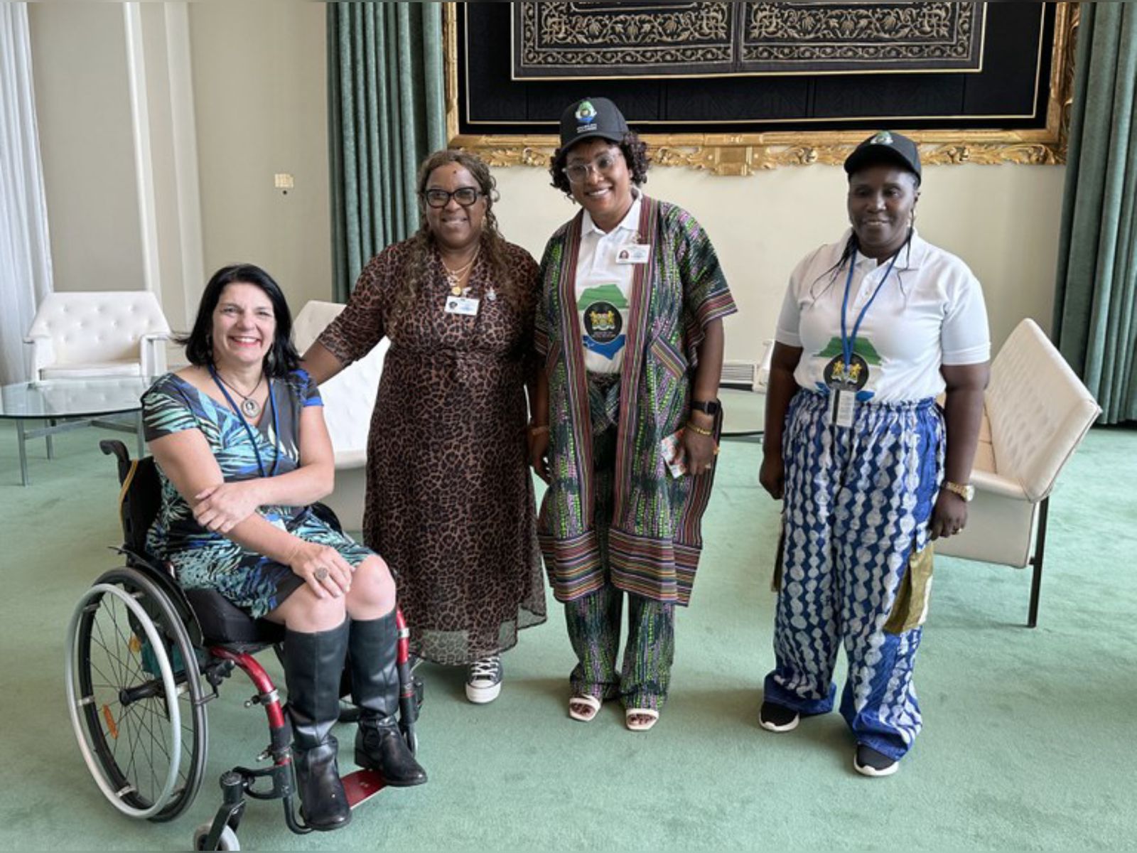 Quatro mulheres posam juntas em um ambiente elegante. Uma delas está em cadeira de rodas, duas usam roupas tradicionais africanas e crachás, e outra veste um vestido de oncinha. Todas parecem sorridentes e o local tem poltronas brancas e decoração sofisticada.