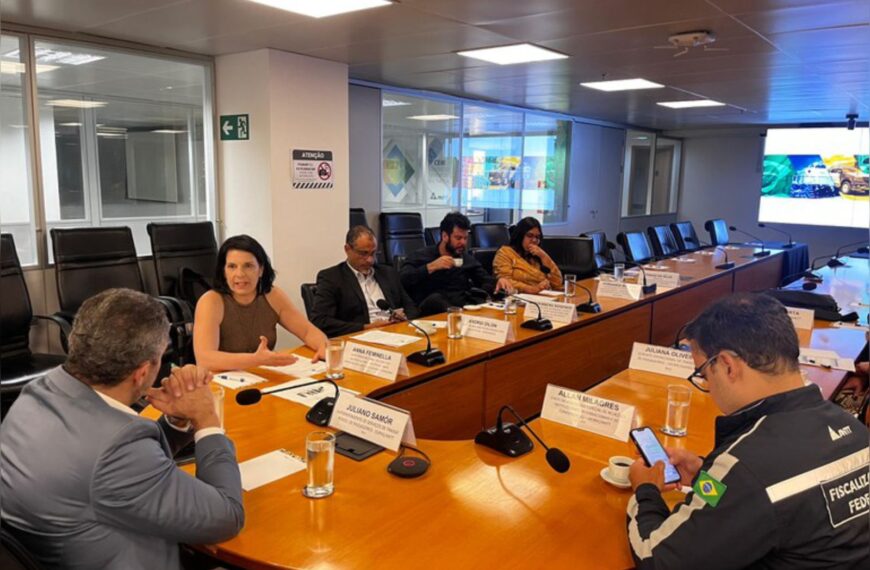 Reunião formal com seis pessoas sentadas à mesa, microfones e crachás de identificação.