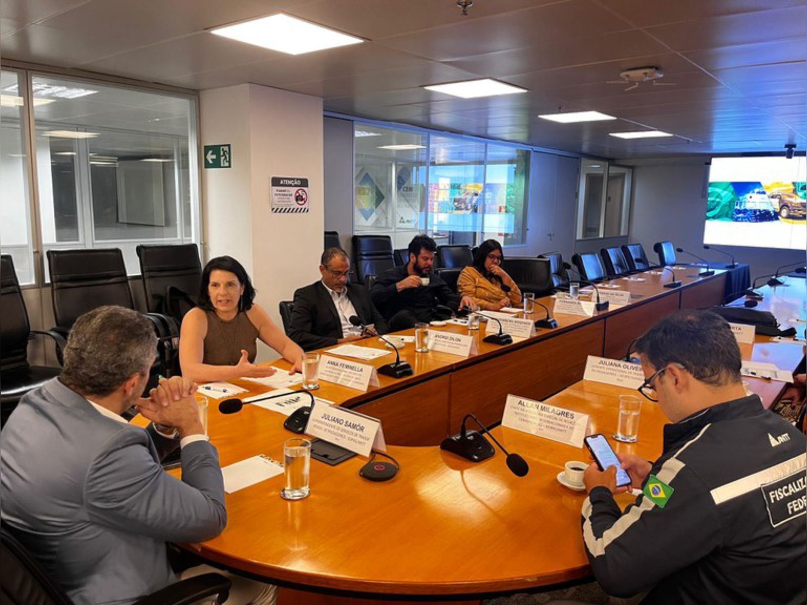 Reunião formal com seis pessoas sentadas à mesa, microfones e crachás de identificação.