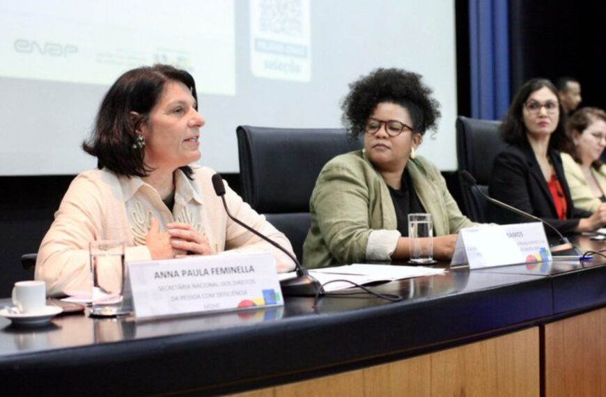 Quatro mulheres estão sentadas em uma mesa de conferência, cada uma com microfone e plaquinha de identificação. A primeira, Anna Paula Feminella, fala ao microfone. O ambiente é formal, com projeção de slides ao fundo.