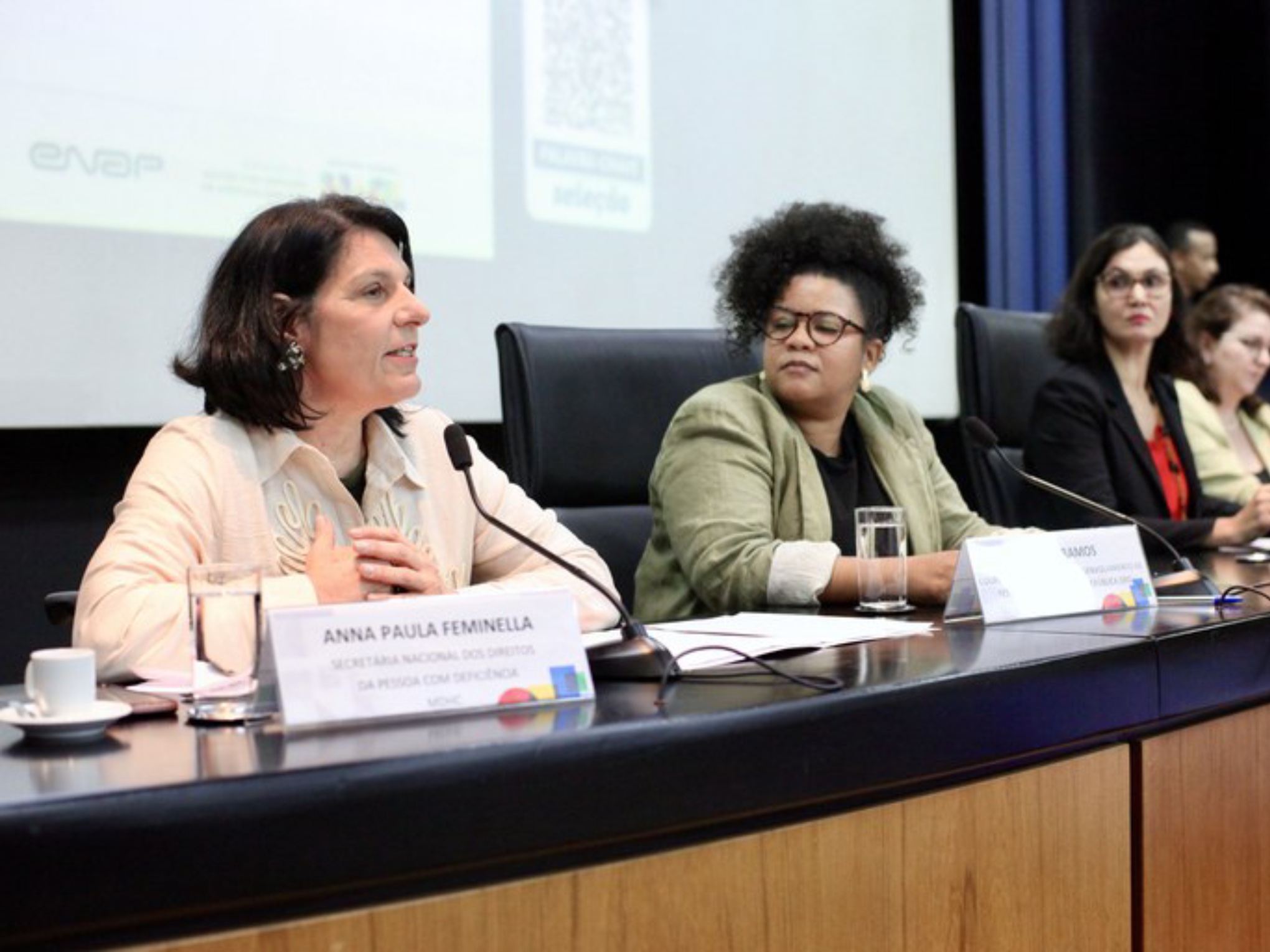 Quatro mulheres estão sentadas em uma mesa de conferência, cada uma com microfone e plaquinha de identificação. A primeira, Anna Paula Feminella, fala ao microfone. O ambiente é formal, com projeção de slides ao fundo.