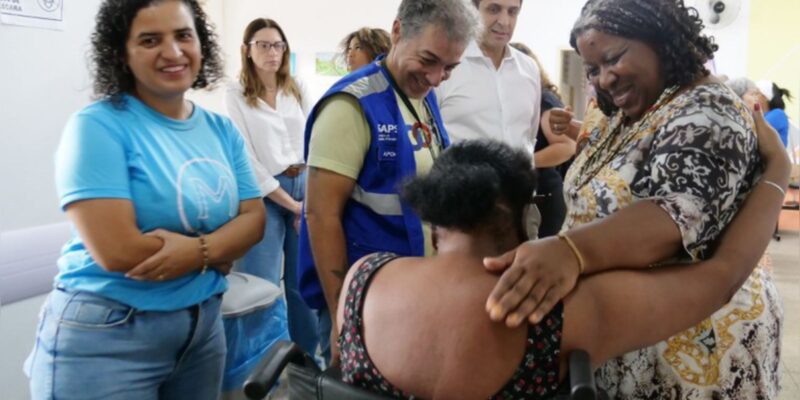 Grupo sorridente em ambiente fechado, com abraço entre duas mulheres, uma em cadeira de rodas.